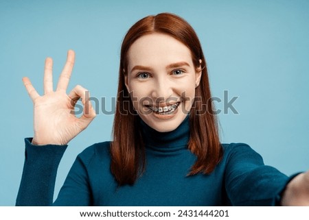 Smiling ginger woman with dental braces showing ok sign looking at camera making selfie isolated on blue background. Positive emotions, health care, orthodontic concept