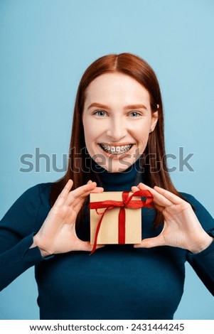 Satisfied young ginger female holding out red wrapped gift box, giving present, looking smiling at camera, wearing casual style clothes. Indoor studio shot isolated on blue background 