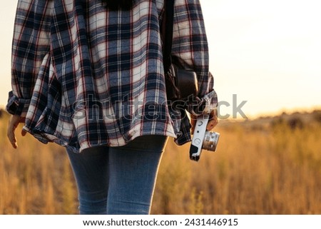 Unrecognizable woman in checkered shirt and casual clothes taking photos with an analog camera in the field at sunset. The image conveys the concept of retro and vintage