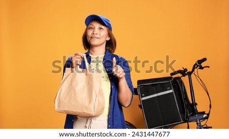 Slow motion portrait shot of friendly courier waiting for customer to answer door, showing thumbs up sign. Food delivery cyclist handing lunch bag to customer, isolated over studio background