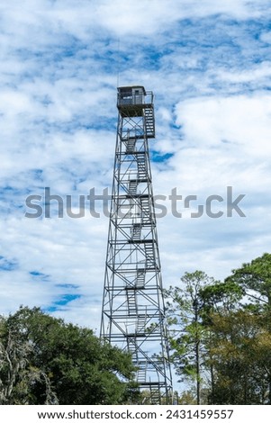 Fire lookout tower high in the sky with tall trees all around. The building has many steps leading up to the tower structure. The small square building has a metal roof and windows on four sides.  Royalty-Free Stock Photo #2431459557