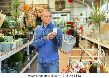 European man picking flower pot and taking pictures with his smartphone while standing in salesroom of store.
