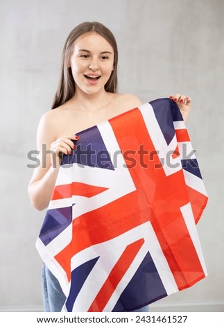 Positive young teen girl in casual clothes holds unfurled flag of Great Britain in hands against gray wall, studio shot. Support, national identification
