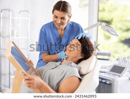 Friendly experienced female aesthetician giving professional advice before hardware facial skin treatments to elderly woman looking in mirror in cosmetological office Royalty-Free Stock Photo #2431462093