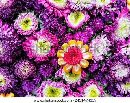 In the picture there are pink and purple carnations, some with white flowers in the middle, arranged together in the center of the picture are orange corn seeds and dark pink amaranth flowers in the m