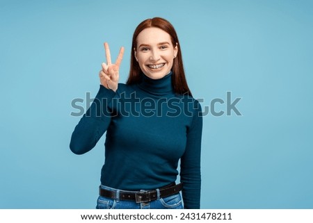 Smiling beautiful redhead with braces, clad in a polo neck, making the peace sign and looking into the camera, isolated on a blue background