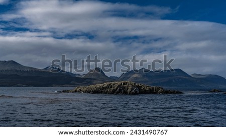 Small rocky islets in the Beagle Channel. Ripples on the blue water. The coastal mountain range of the Andes against the background of sky and clouds. Tierra del Fuego Archipelago. Argentina