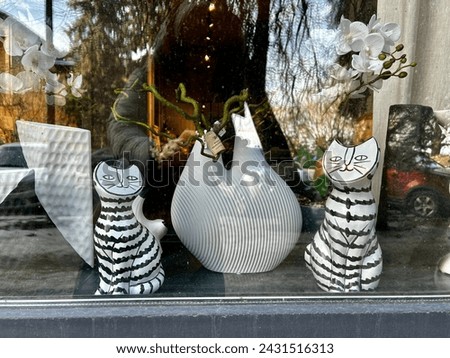 horizontal photo with a view of a window decorated with plaster figures of striped cats, near a large vase with orchids on the windowsill. 