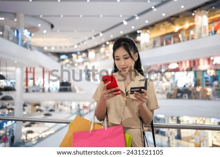 young woman in smart casual wear carrying many paper shopping bags shopping online on mobile phone using credit card at shopping center Royalty-Free Stock Photo #2431521105