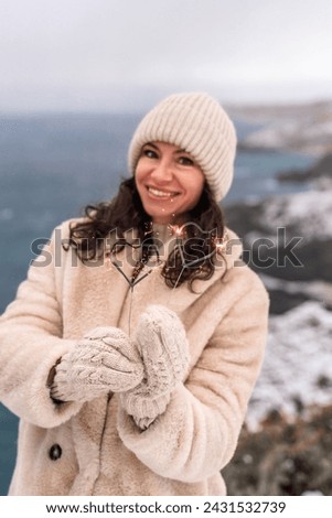 Outdoor winter portrait of happy smiling woman, light faux fur coat holding heart sparkler, posing against sea and snow background