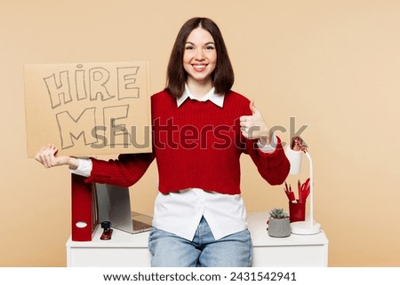 Young employee business woman wear red sweater shirt work at office sit on desk with pc laptop hold cardboard sign card hire me isolated on plain pastel beige background. Achievement career concept