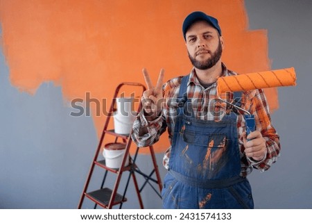 Painter man in overalls with painting roller smiling and showing victory sign. Close-up