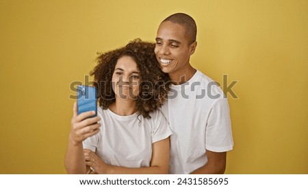 Confident beautiful couple, oozing joy, taking a smiling selfie on their smartphone. standing together, they create a fun expression against isolated yellow background