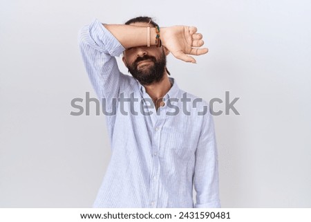 Hispanic man with beard wearing casual shirt covering eyes with arm, looking serious and sad. sightless, hiding and rejection concept  Royalty-Free Stock Photo #2431590481