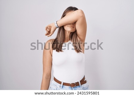 Hispanic young woman standing over white background covering eyes with arm, looking serious and sad. sightless, hiding and rejection concept  Royalty-Free Stock Photo #2431595801