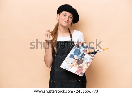 Young artist caucasian woman holding a palette isolated on beige background making money gesture