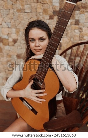 A woman holding an acoustic guitar on a wooden chair in front of a brick wall