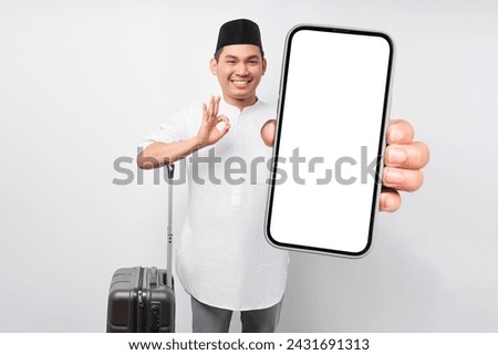 cheerful young Asian Muslim man standing while holding a suitcase and smartphone, showing ok okay gesture isolated on white background. Ramadan and eid Mubarak concept