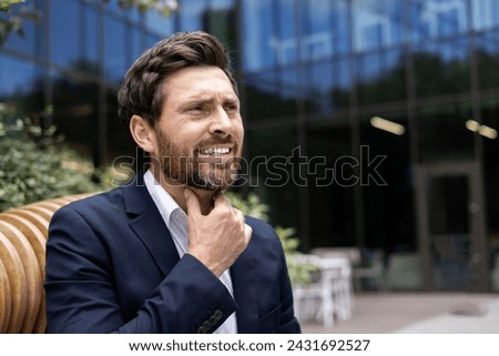 A sick young man in a business suit sits on a bench on the street and holds his hand to his throat, feels severe pain. Close-up photo.
