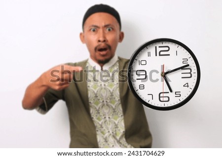 A shocked Indonesian Muslim man in koko and peci emphatically points at a clock, realizing he's late for his sahur meal during Ramadan. Isolated on a white background