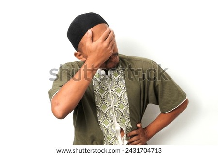 Embarrassed Indonesian Muslim man in koko shirt and peci covers his face in a facepalm. Isolated on White background
