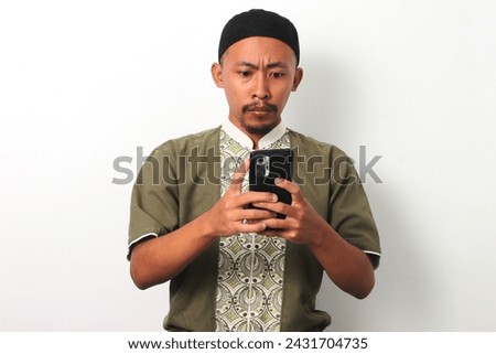 Shocked Indonesian Muslim man in koko and peci reacts with surprise or concern to something on his phone during Ramadan. Isolated on a white background