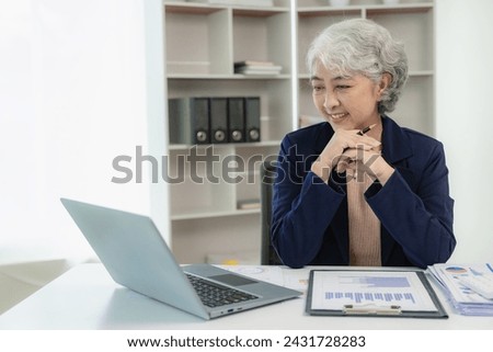 Senior Asian businesswoman working with graph documents to analyze finances and work sitting at table in office, typing on keyboard, studying financial documents, analyzing profit margins.