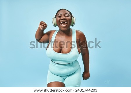 cheerful plus size african american woman dancing in active wear with headphones on, blue background