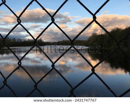 A sunset reflection on a calm lake behind a chainlink fence. Royalty-Free Stock Photo #2431747767