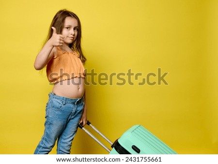 Caucasian adorable little kid, tourist traveler girl in summer clothes, girl thumbing up looking at camera, carrying plastic trendy suitcase, going for weekend getaway, isolated yellow studio backdrop