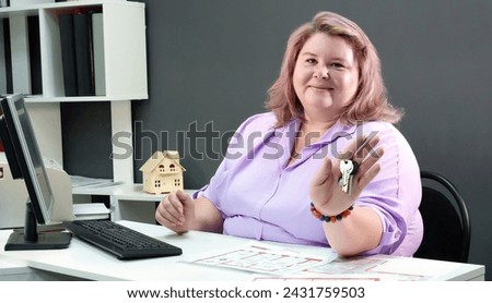 middle-aged woman shows a bunch of keys while, sitting at a table in front of a computer against a gray wall in her office.