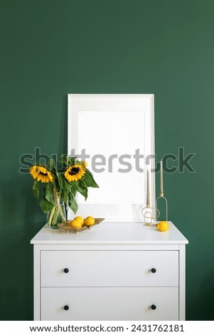 Vertical shot of white commode with blank picture in frame, candlestick, plate with lemons and yellow sunflowers in glass vase on green wall background in modern living room.