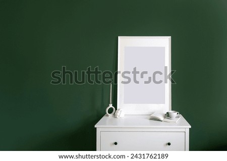 Candle, open book, ceramic cup and empty picture in frame on white drawer dresser in living room on green wall background with copy space. Home decor idea.
