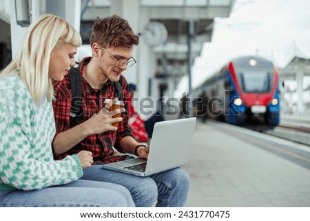 A young traveler couple is sitting on a train station and using a laptop. Passengers are waiting for train departure and typing on a laptop while smiling at it. Young commuters on railway station. Royalty-Free Stock Photo #2431770475