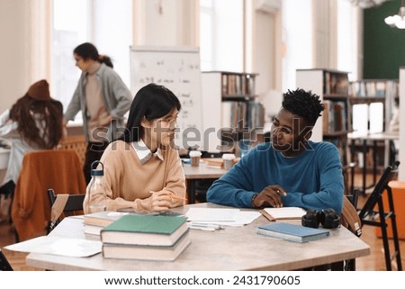 Portrait of young Asian woman and Black man sitting at table in class together during pair study Royalty-Free Stock Photo #2431790605