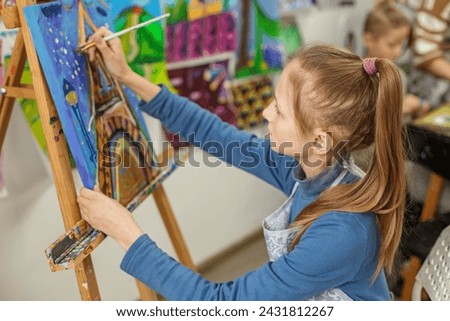 Young girl with ponytail, deeply concentrated while painting colorful image on canvas during an art class