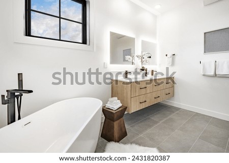Primary bathroom interior featuring grey slate tile white walls free standing bathtub floating cabinet sink decor large walk in shower windows with black window frame small rug on the floor Royalty-Free Stock Photo #2431820387