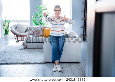 Full body photo of charming nice aged lady sit sofa hold house key demonstrate thumb up approval spacious interior indoors