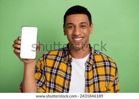 Photo of young student smiling guy in checkered shirt demonstrate smartphone display tap touchscreen isolated on green color background