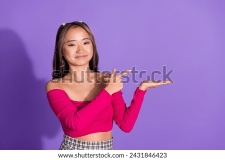 Photo of adorable cheerful girl with ponytails wear stylish crop top indicating at offer on palm empty space isolated on violet background