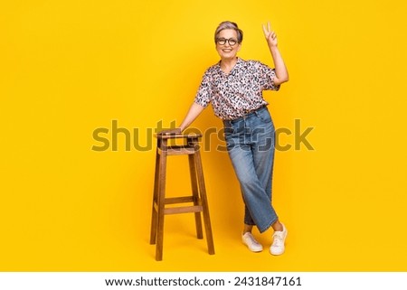Full size photo of pretty retired female lean on chair show v-sign wear trendy leopard print outfit isolated on yellow color background