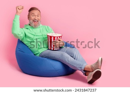 Full length photo of handsome senior man sit bean bag winning eat popcorn dressed stylish green garment isolated on pink color background