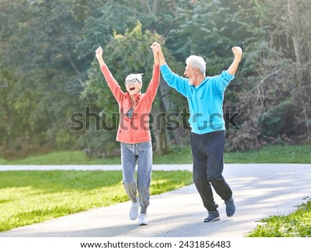 Smiling active senior couple jogging exercising and having fun and celebrating success rasing hands together taking a break in the park Royalty-Free Stock Photo #2431856483