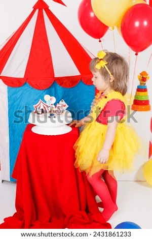 A little girl in a colorful outfit is looking at the birthday cake. Circus theme. White, blue, red and yellow colors. Vertical image. 