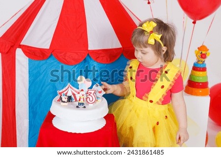 A little girl in a colorful outfit is looking at the birthday cake. Circus theme, white, blue, red, yellow colors. Horizontal picture. 