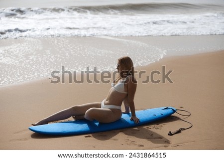 Young beautiful blonde surfer woman in white bikini sitting on blue surfboard. The girl enjoys her holidays on the beach to practice her favourite sport. Travel and holiday concept