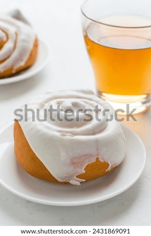 Two buns covered with white glaze on a saucer with a cup of tea on a light background.