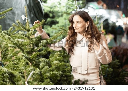 Positive interested young woman with wavy brown hair looking for freshly cut Christmas tree on outdoor market..