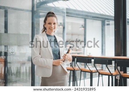 Business management concept, A smiling young professional woman stands in a modern office, holding a tablet, ready for a productive workday. Royalty-Free Stock Photo #2431931895