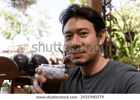 Happy funny face Asian man selfie himself drinks fresh iced coffee in the cafe garden.